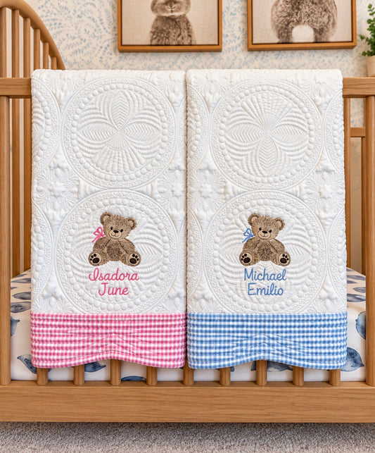 Two personalized teddy bear baby quilts with pink and light blue gingham trim hanging on a wooden crib