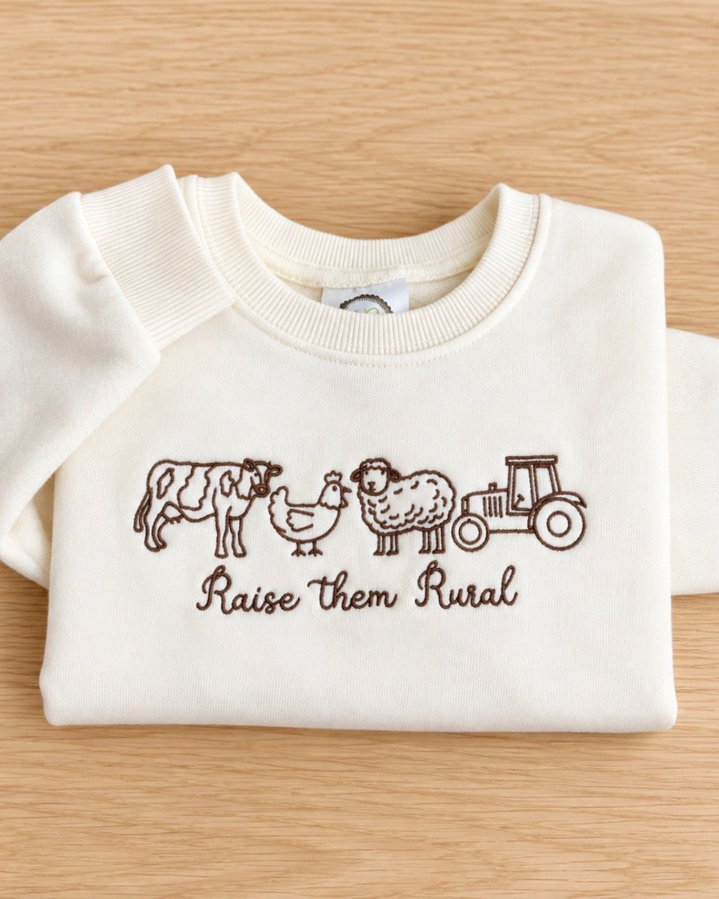 Cream toddler sweatshirt embroidered with a cow, chicken, sheep and tractor above Raise them Rural