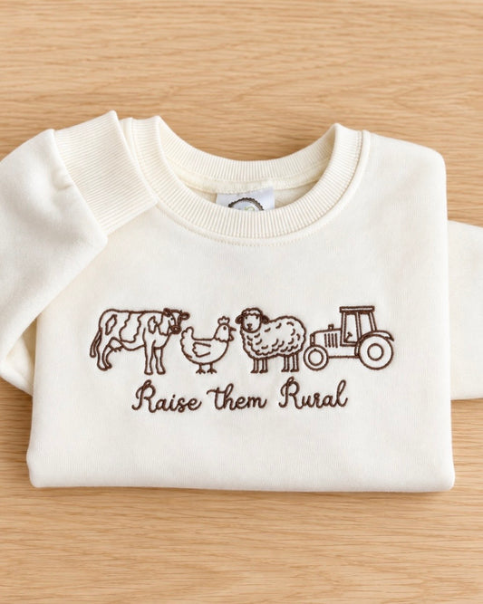 Cream toddler sweatshirt embroidered with a cow, chicken, sheep and tractor above Raise them Rural